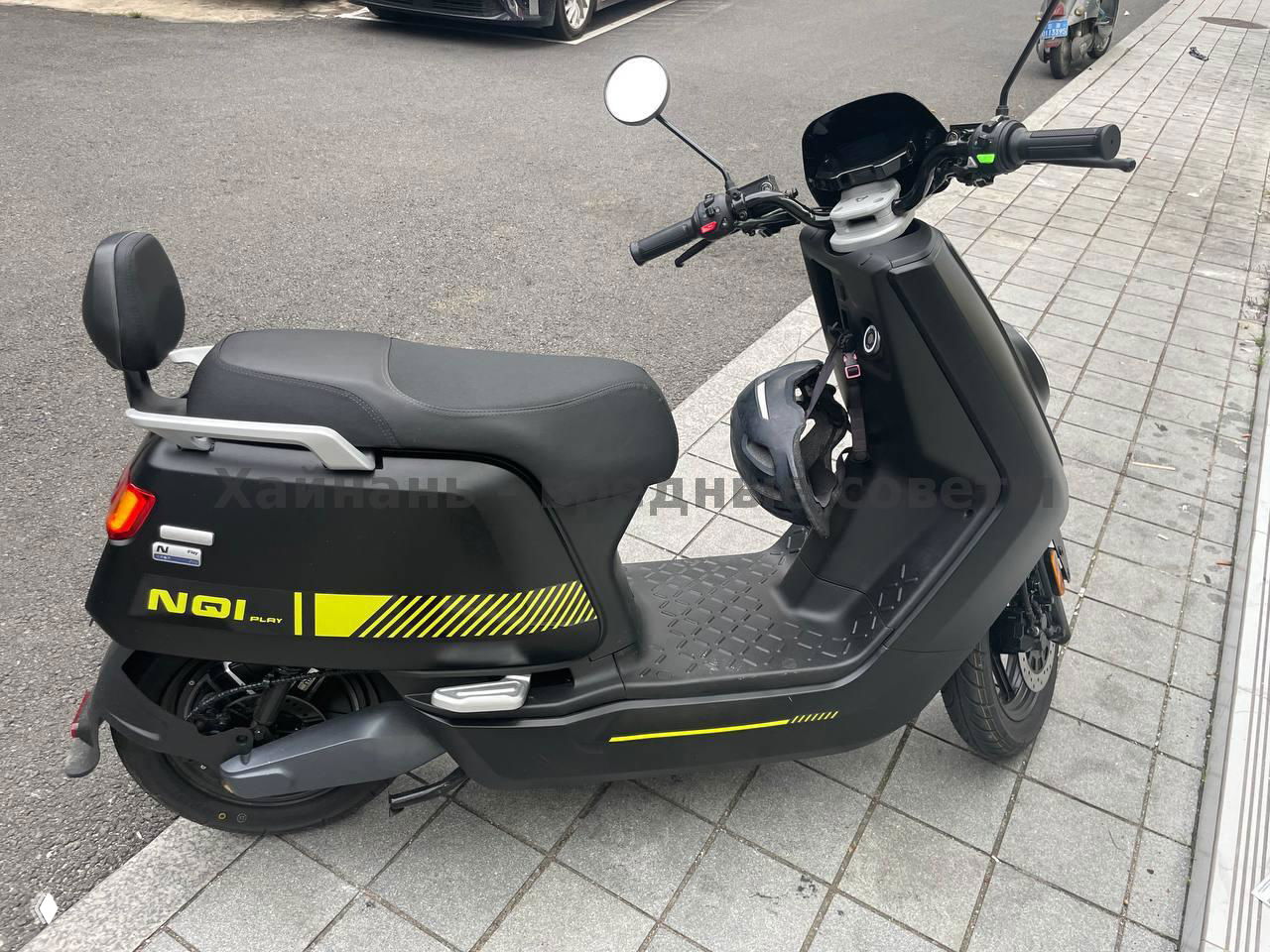 Черный электрический скутер NIU с жёлтыми декоративными полосами, боковой ракурс на плиточном тротуаре рядом с магазином в Санье, виден шлем и подставка.