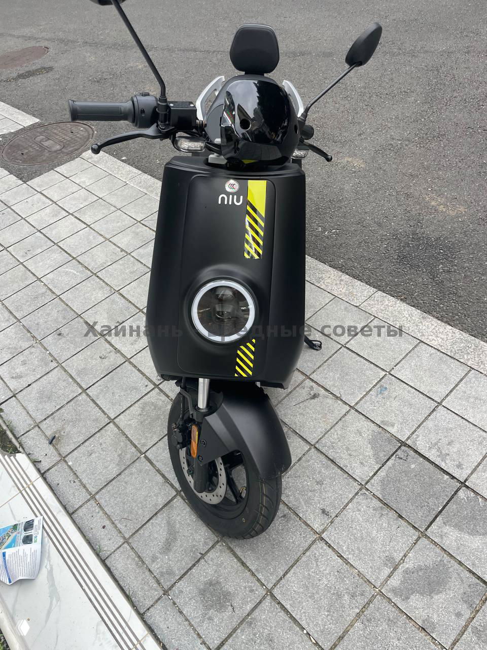Вид спереди на черный скутер NIU с круглой фарой и жёлтой полосой по капоту, шлем на руле, скутер припаркован на тротуаре в городском окружении.