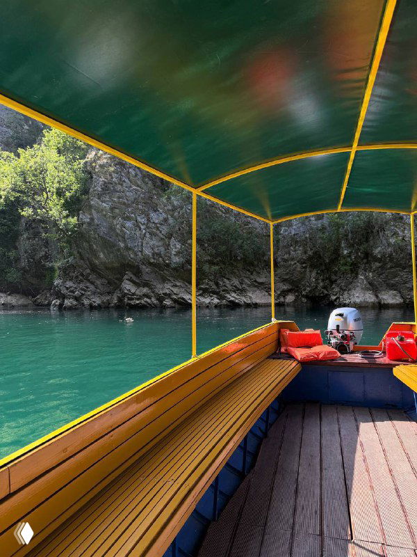 Вид с крытой лодки: деревянные скамьи и зелёный тент, отражение скал в воде и туристическое судно на реке.