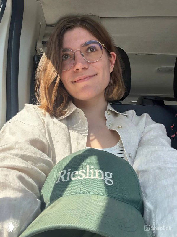 Портрет автора в машине: женщина в очках и светлой рубашке улыбается, на кепке надпись Riesling, дорожный кадр.
