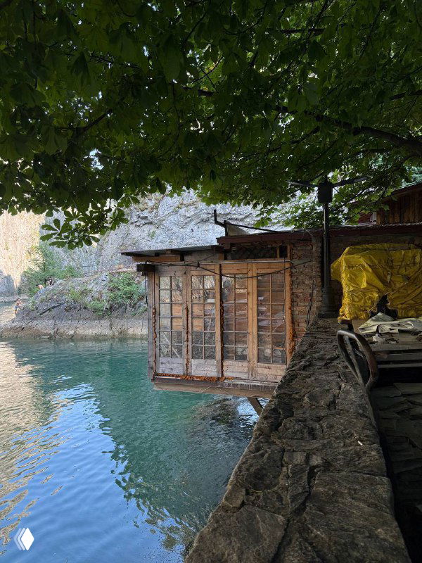 Небольшая деревянная пристань и домик у скалы под навесом, спокойная вода и тень от листвы в природном резервате.