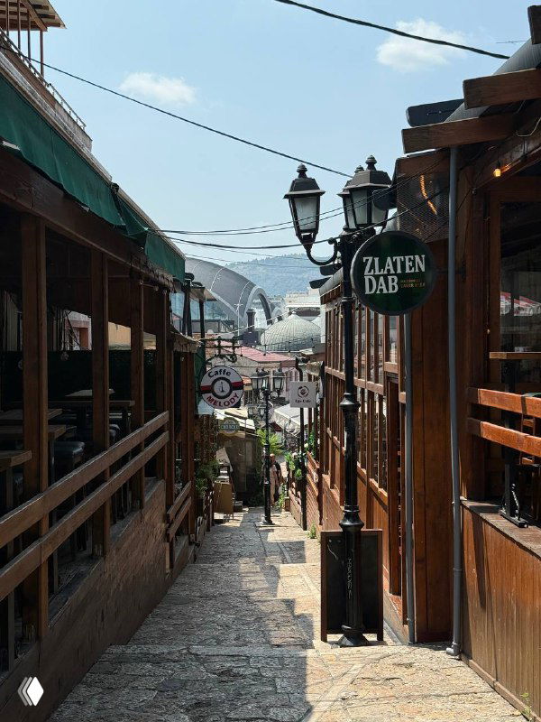 Узкая пешеходная улица в старом городе с деревянными лавками и вывесками, туристическая атмосфера и вид на холмы.