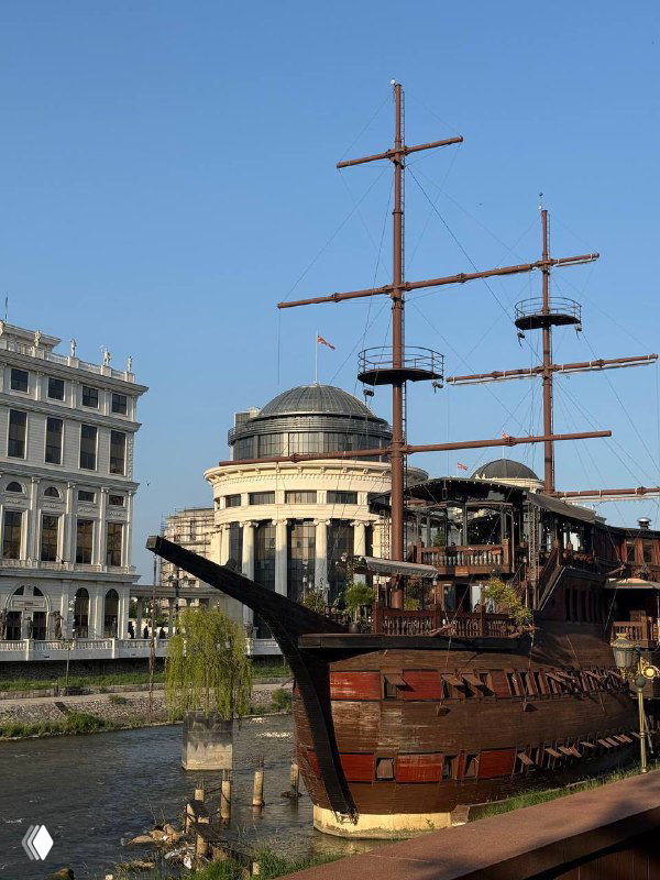 Деревянный корабль-реплика у каменной набережной на фоне городской застройки и купола, туристический объект на берегу.