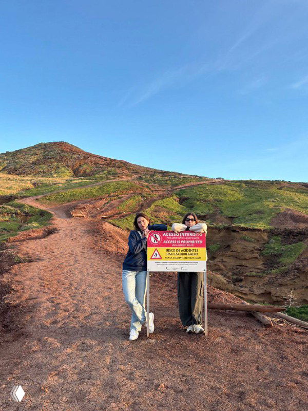 Две женщины стоят на красно-земляной тропе у предупреждающего знака на фоне холмов и синего неба, сценка из прогулки по природной локации.