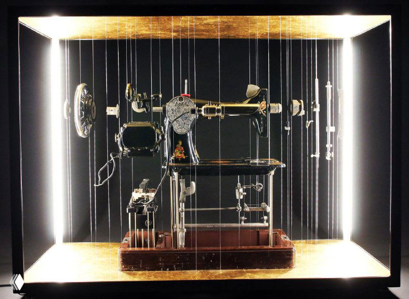 Инсталляция с разобранным механическим прибором: многочисленные металлические детали подвешены в витрине, создавая эффект взрывной сборки и драматичную подсветку.