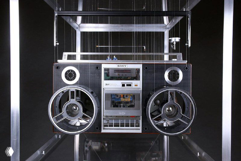 Инсталляция из разобранной аудиотехники: магнитофон и динамики демонстрируют внутреннее устройство устройства, элементы висят на нитях в прозрачном коробе.