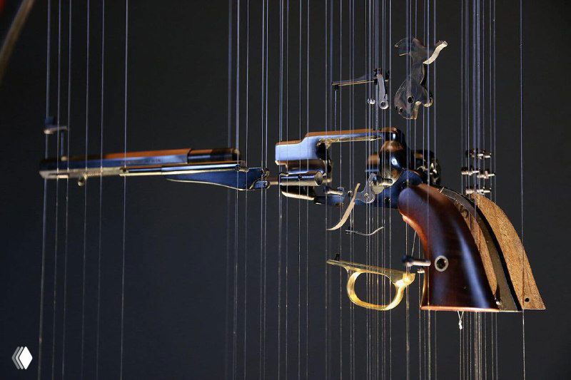 Художественная инсталляция с разобранным огнестрельным элементом: части пистолета аккуратно подвешены в пространстве как объект дизайна и механики.