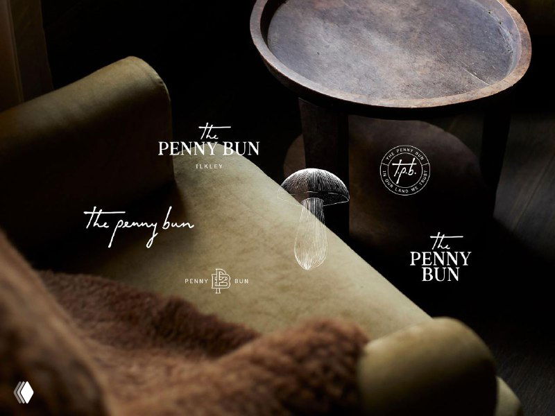 Несколько вариантов логотипа The Penny Bun на уютной интерьерной сцене: кружки, ткань и графические эмблемы бренда.