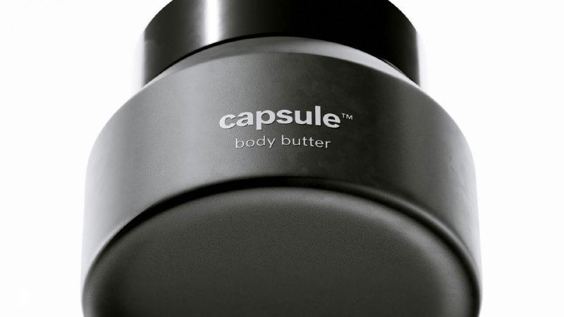 Крупный план чёрной баночки с надписью «capsule» на минималистичном сером фоне; акцент на форме и материале упаковки для ухода за кожей.