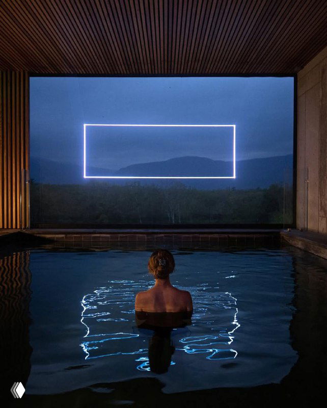 Человек в воде смотрит на прямоугольную неоновую рамку над ландшафтом — кадр, объединяющий воду, свет и окружающий пейзаж в инсталляции.