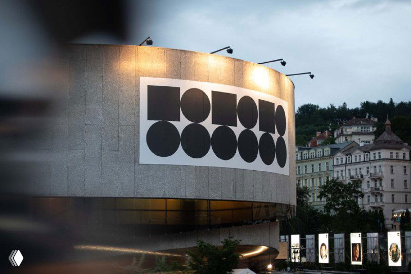 Фасад здания фестиваля в Карловых Варах с крупным черно‑белым модульным логотипом из кругов на бетонной поверхности вечером.