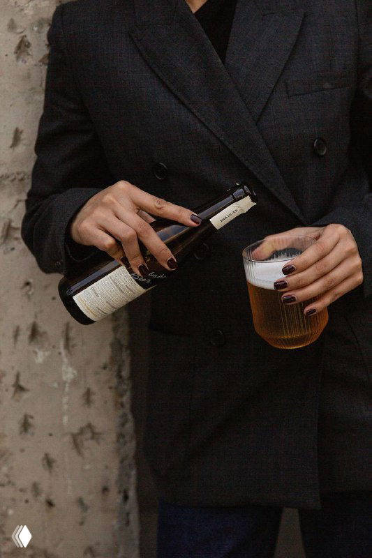 Сцена: человек в тёмном пиджаке наливает комбучу Brain+er в стакан; видны бутылка с утилитарной этикеткой, белый леттеринг и рука с маникюром.