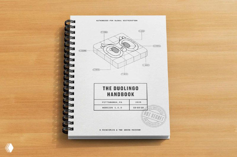 Спиральный блокнот на деревянном столе с обложкой «The Duolingo Handbook», схематичным рисунком и оттиском печати — макет корпоративного руководства.