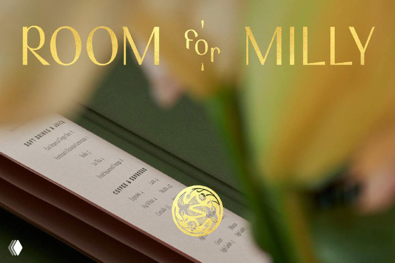 Крупный кадр разворота меню с золотым логотипом «ROOM for MILLY», мягкое переднее размытие и цветочный план на переднем фоне — художественная подача айдентики.