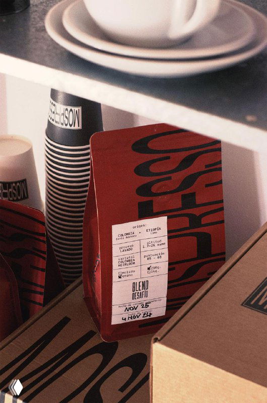 Красный пакет Mospresso в коробке на полке рядом с бумажными стаканчиками; демонстрация минималистичной упаковки, крупной утилитарной типографики и композиции.