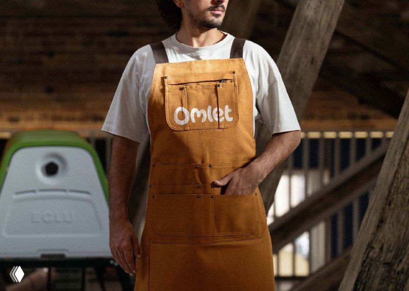 Фотография мужчины в фартуке с логотипом Omlet на фоне мастерской и видимым продуктом бренда — пример фирменной одежды и предметной стилизации