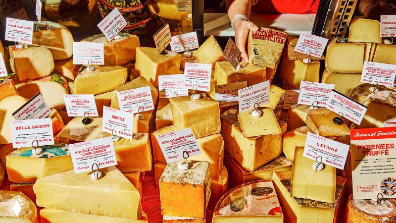 Прилавок с множеством кусков сыра и мелкими ценниками; изображение иллюстрирует ассортимент и визуальную айдентику сети Murray’s в торговом интерьере.