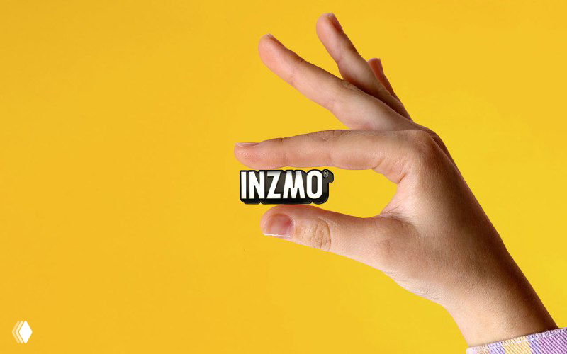 Рука держит стикер или элемент фирменной айдентики с логотипом Inzmo на жёлтом фоне — пример использования знака в материалах бренда.