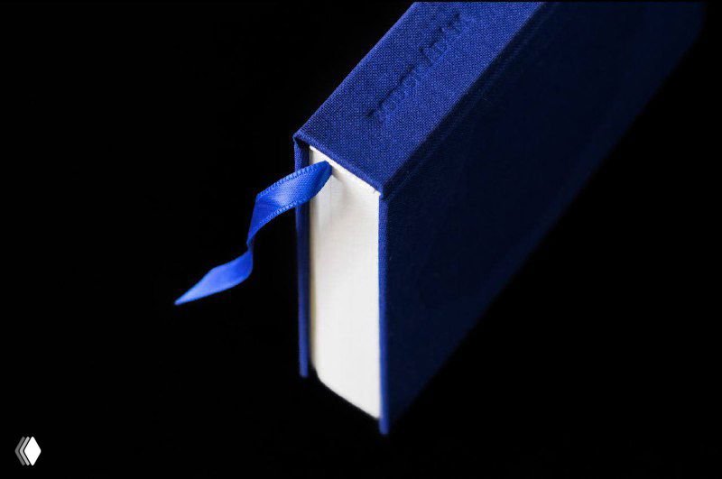 Крупный план синего переплёта книги с лентой-закладкой на тёмном фоне: виден корешок, плотный переплёт и аккуратная отделка краёв из ткани