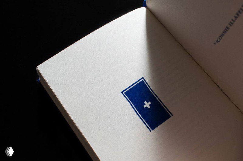 Крупный план внутренней печатной вставки: синий прямоугольник с белым символом на текстурной бумаге, подчёркивающий графическую идею редизайна