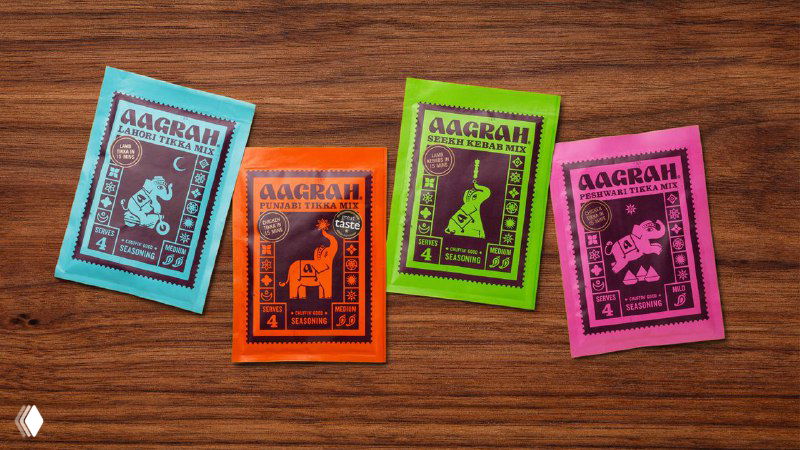 Разноцветные саше Aagrah на деревянной поверхности: яркие монохромные упаковки с орнаментами и игривыми изображениями слонов и индийских мотивов.