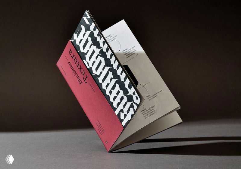 Один экземпляр зина «Blackletter. Textura» в развороте: красная обложка и чёрно‑белый узор текстуры; игра света и тени подчёркивает форму.