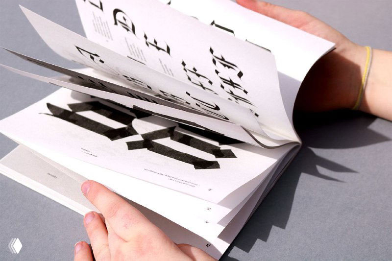 Человек перелистывает страницы зина «Blackletter. Textura»: чёрные каллиграфические мотивы и крупные графические формы на белом фоне.