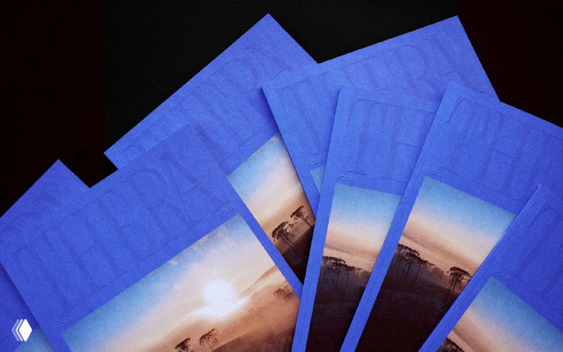 Несколько экземпляров журнала Thera Mag с насыщенно‑синей тиснёной обложкой, разложенных веером; видна вставка с приглушённой фотографией пейзажа.