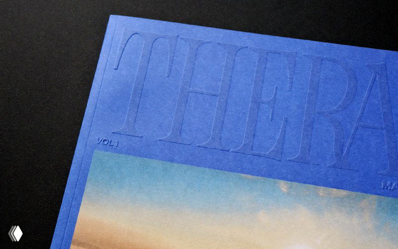 Крупный план синей обложки Thera Mag: заметно тиснёное название и нижняя вставка с фотографией пейзажа в контрасте с текстурой бумаги.