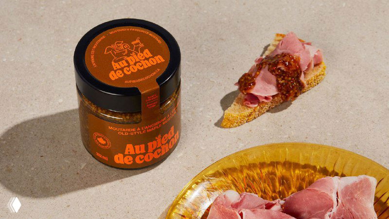 Банка с этикеткой Au Pied de Cochon рядом с тостом и ломтиком ветчины на пастельном фоне; упаковка в тёплых бежево-оранжевых тонах.