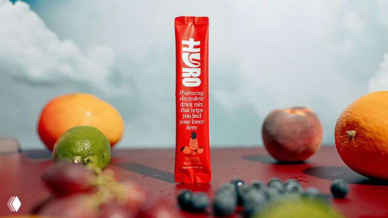 Вертикальное саше Hyro на столе на фоне фруктов и неба; контраст красной упаковки и белого логотипа с пастельными элементами композиции.