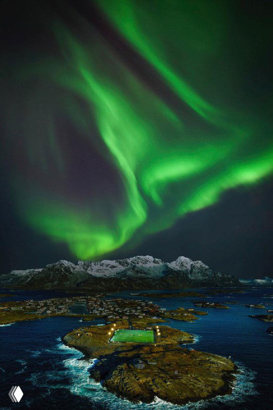 Полярное сияние над скалистым побережьем: зелёные волны в небе, освещающее океан и небольшие острова, снято с высоты дрона.
