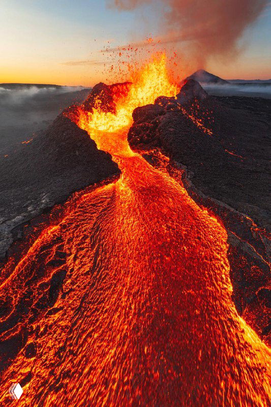 Активный вулкан с лавовым потоком: яркие оранжево-красные потоки лавы контрастируют с тёмной поверхностью окрестностей, снято сверху.