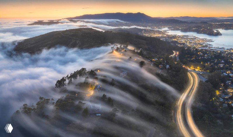 Туман, стекающий по холмам, и извилистая дорога с автомобильными огнями: вид сверху показывает динамику облаков и ночную подсветку ландшафта.