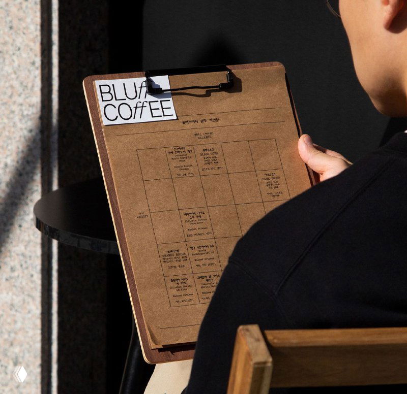 Бариста держит планшет с меню или рабочим графиком; видна фирменная наклейка Bluff Coffee — пример брендированных материалов и упаковки.