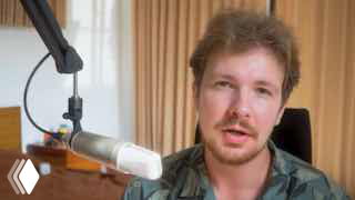 Крупный план ведущего (Олег Стефанов) за столом с конденсаторным микрофоном; на заднем плане шторы и рабочая/студийная обстановка в помещении.