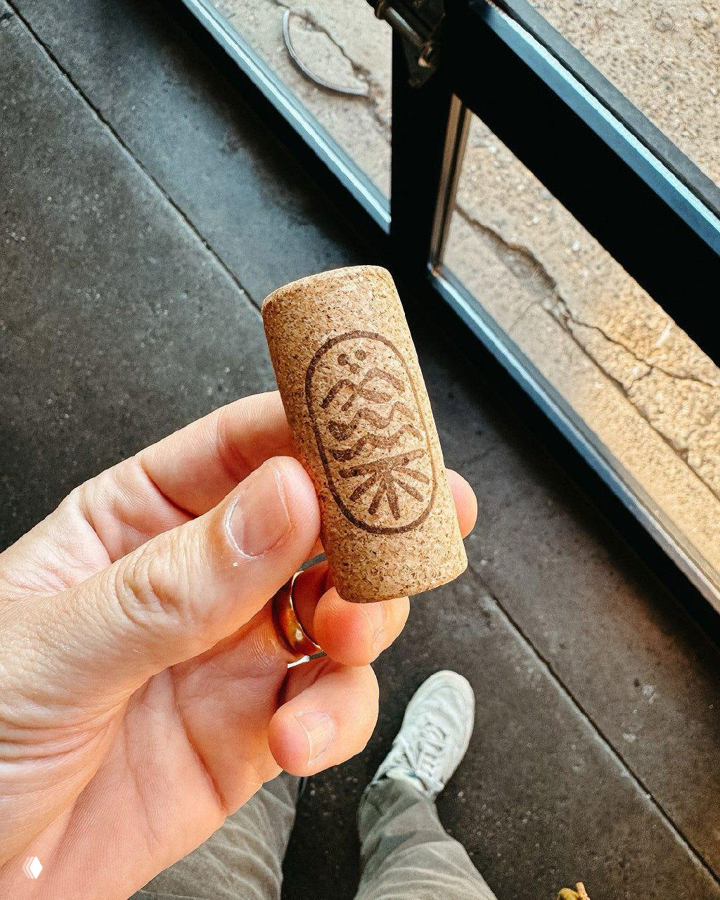 Рука держит винную пробку с тиснением логотипа Living Roots на фоне пола и стеклянной двери — деталь крафтовой айдентики бренда.