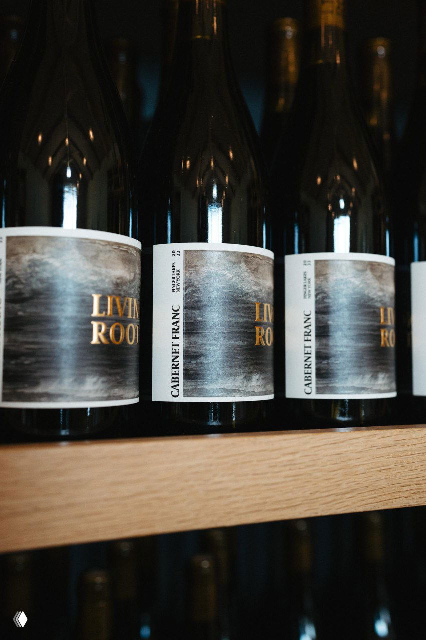 Ряд бутылок Cabernet Franc с тёмными этикетками Living Roots на деревянной полке — магазинная выкладка и визуальное позиционирование продукта.