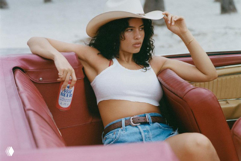 Женщина в шляпе и лёгкой одежде держит банку «Not Beer» в салоне ретро‑автомобиля; кадр подчёркивает упаковочный дизайн и атмосферу съёмки