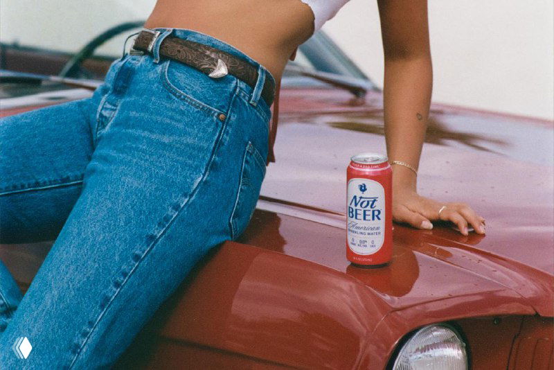 Кадр с моделью, сидящей на капоте автомобиля рядом с банкой «Not Beer»; акцент на дизайне банки и визуальном стиле рекламной серии фотографий