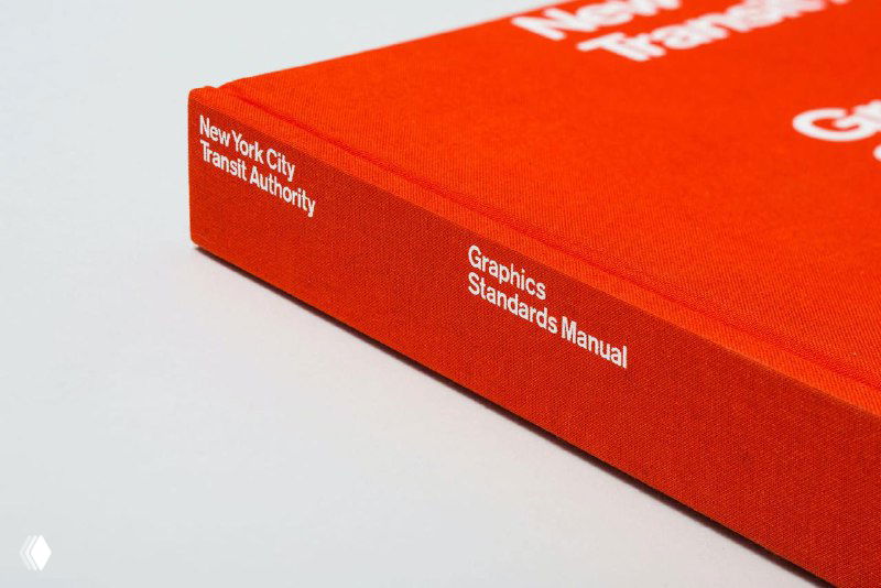 Крупный план красного переплёта переиздания NYCTA Graphics Standards Manual на светлом фоне, видна тисненая надпись на корешке книги.