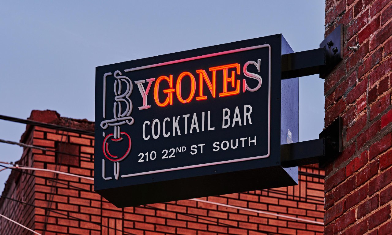 Неоновая вывеска Bygones Cocktail Bar на фасаде кирпичного здания вечером; ретро-стиль вывески и видимая строка адреса на табличке.