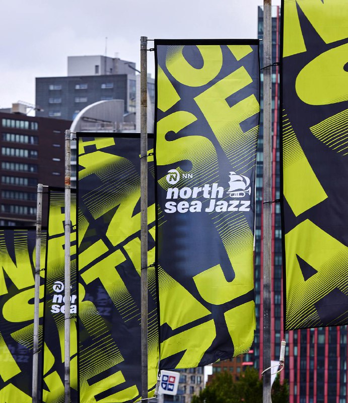 Уличные флаги с ярко‑лимонной типографикой North Sea Jazz на вертикальных баннерах вдоль городской улицы, пример брендирования общественного пространства и событийной навигации.