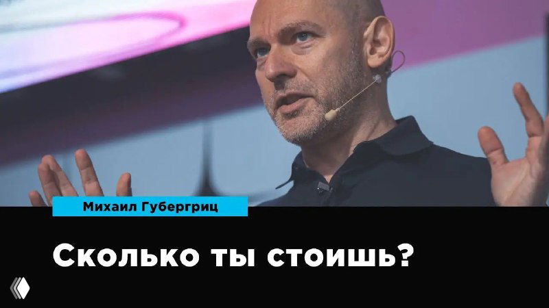 Михаил Губергриц во время лекции: крупный план спикера, на изображении видна надпись «Сколько ты стоишь?» на тёмном баннере.