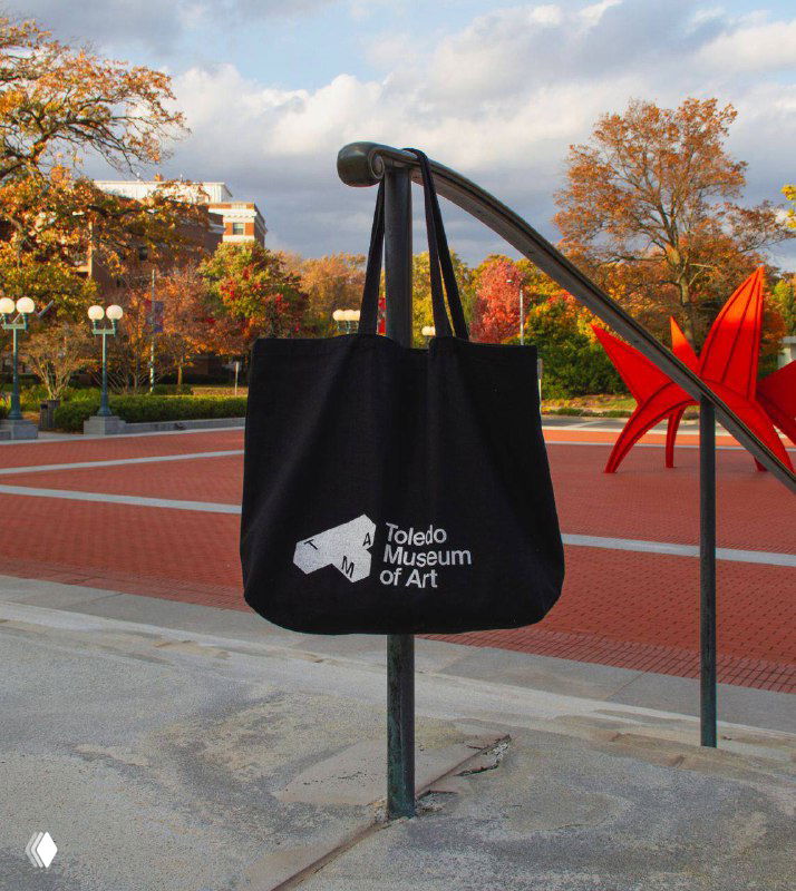 Чёрная тканевая сумка с белым логотипом Toledo Museum of Art, подвешенная на уличной стойке; пример сувенирного мерча и брендирования аксессуаров.