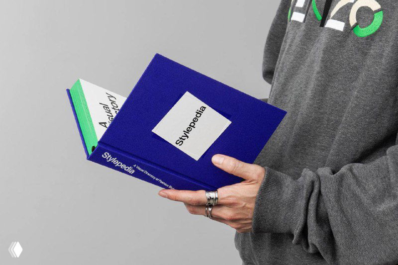 Руки человека держат две копии «Stylepedia»: макет демонстрирует масштаб, фактуру текстиля обложки и яркие зелёные торцы страниц.