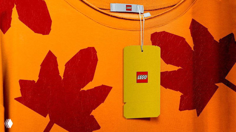 Фотография бирки на текстильном ярко-оранжевом изделии с маленьким логотипом LEGO, показывающая применение фирменных элементов на мерче.