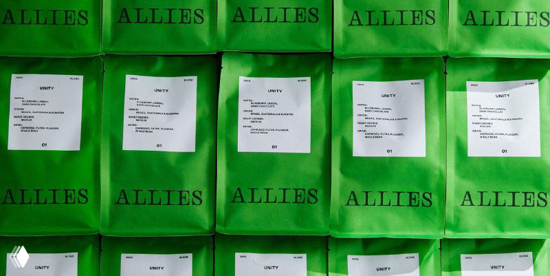 Ряд зелёных пакетов с логотипом ALLIES и белыми наклейками на текстурированном фоне — крупный план фирменной упаковки кофейного бренда в минималистичном визуальном стиле