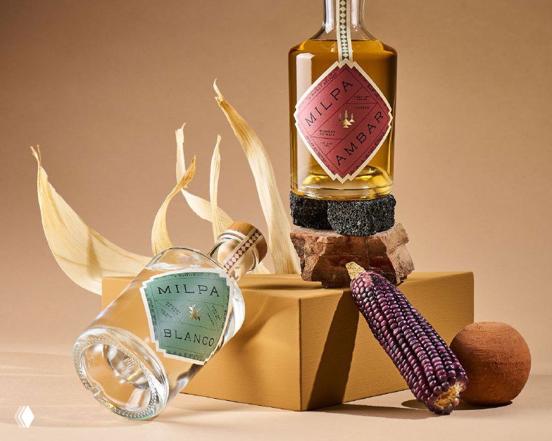 Бутылка Milpa на подиуме рядом с декоративной кукурузой и керамическими элементами; композиция в тёплых тонах демонстрирует продукт и этикетку.