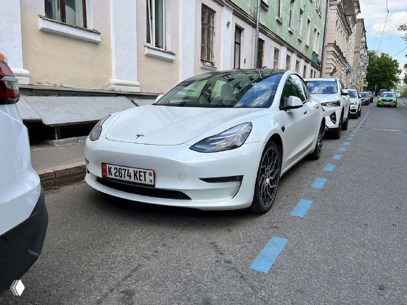 Электромобиль Tesla на городской улице с экспериментальным вариантом госномера, вид спереди-сбоку, голубая разметка и дома на фоне.